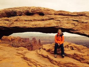 Marlo Bennett in orange jacket sitting at canyon's edge under a stone arh